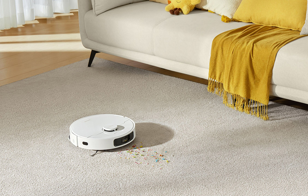 biały robot sprzątający czyści dywan, w tle jasna kanapa i żółty koc